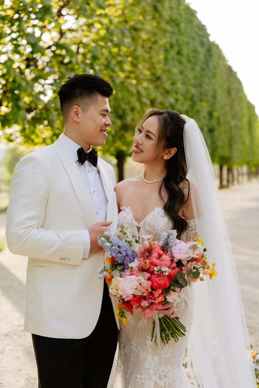 Bridal couple stands on a sunlit Paris garden path, smiling and holding a colorful bouquet