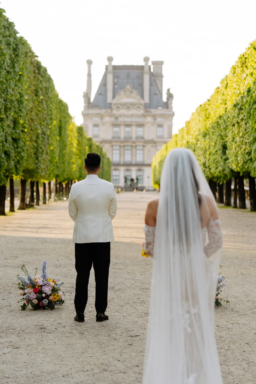 Bride and groom stand facing a grand chateau in a formal garden, with bouquets on the path