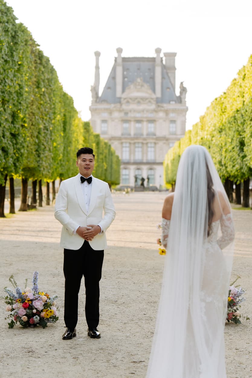 Bride and groom face each other in a tree-lined garden, with floral arrangements and soft daylight