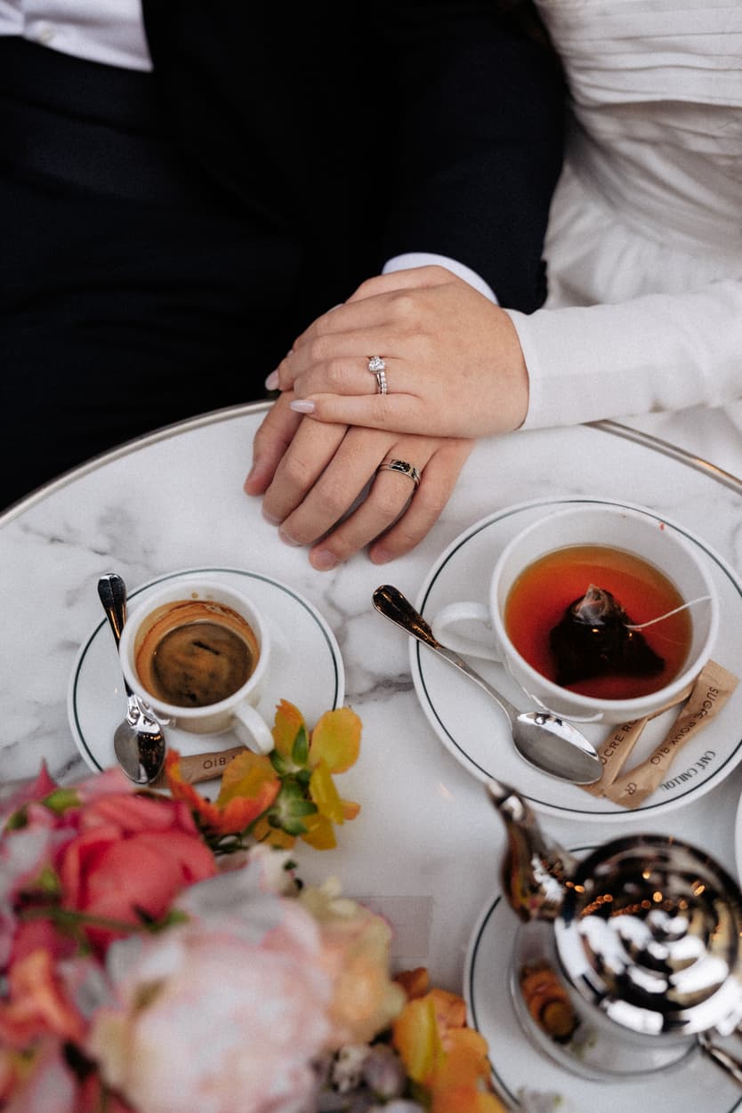 Bride and groom clasp hands over tea cups and flowers at a marble café table in soft light