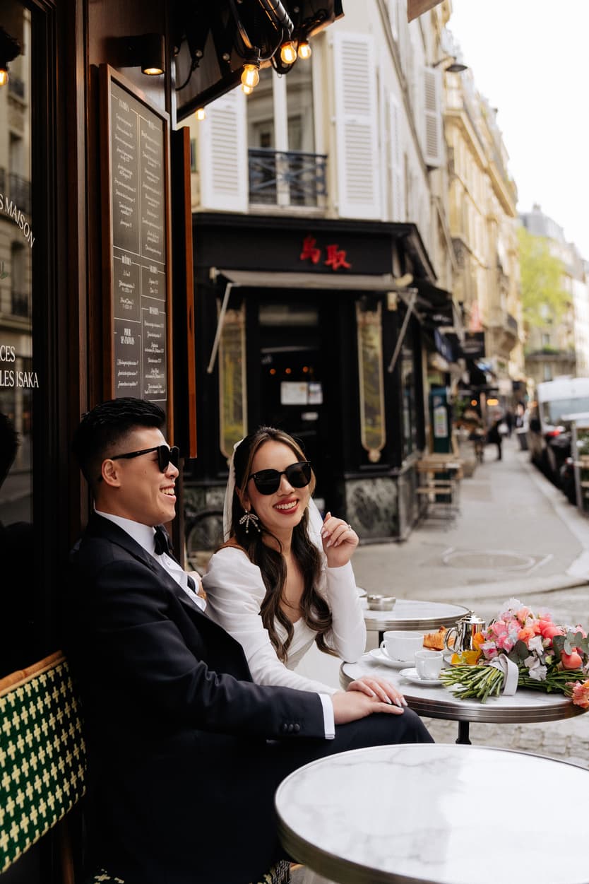 Bride and groom in sunglasses sit at a café table on a Paris street with a bouquet and soft daylight