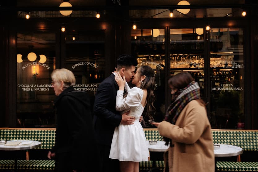Bride and groom kiss outside a Paris café as pedestrians pass in warm evening light