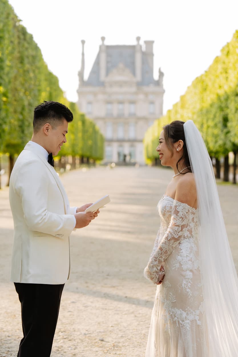 Bride and groom face each other on a tree-lined garden path, with a sunlit historic Paris building behind them