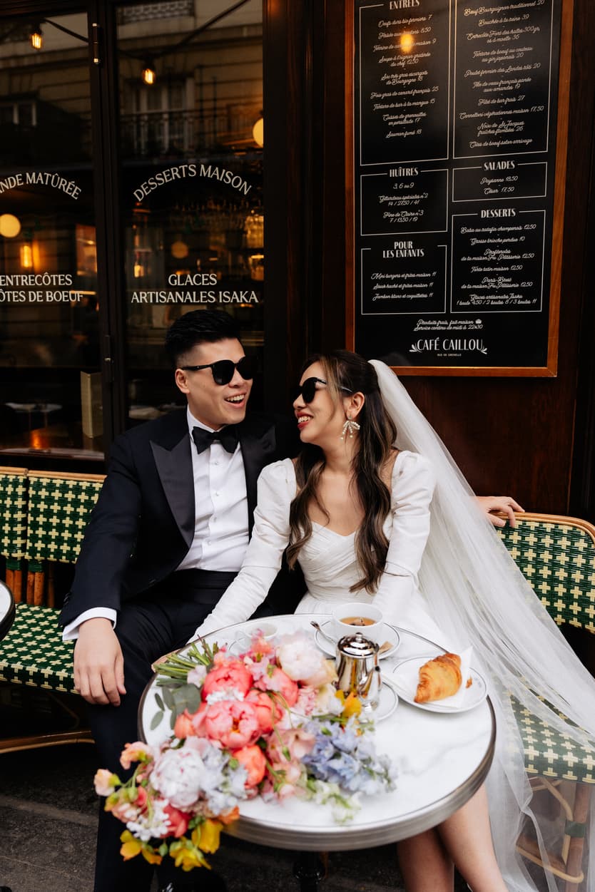 Bride and groom in sunglasses sit at a Paris café table with flowers, pastries and warm window light