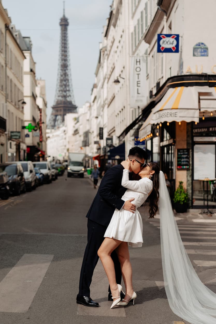 Bride and groom kiss on a Paris street with the Eiffel Tower rising behind them in soft daylight