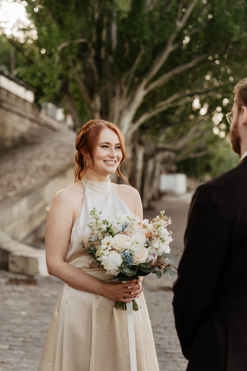 Bride smiles with bouquet at groom on a tree-lined Paris street, soft evening light glowing behind her