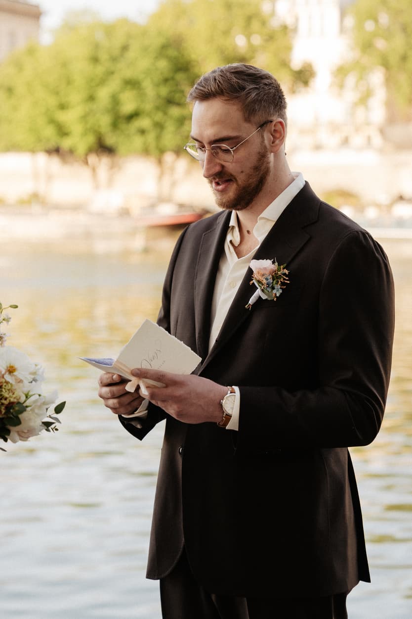 Groom in a black suit reads vows beside the Seine in warm light, holding a small card with a boutonniere