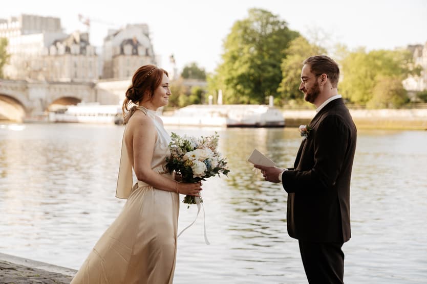 Bride and groom face each other by the Seine in soft light, bouquet in hand