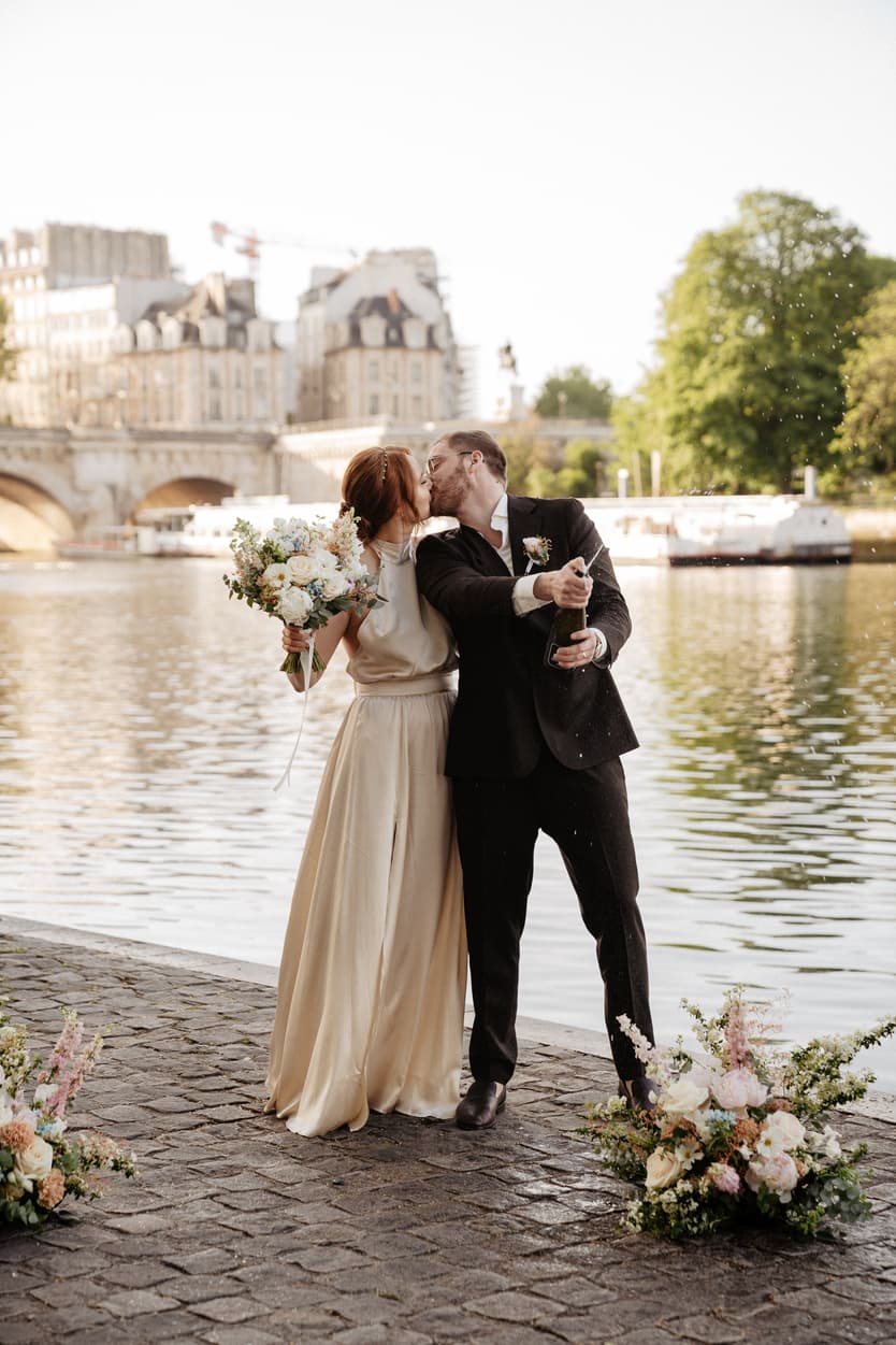Bride and groom kiss on a Paris riverbank as he pops champagne beside floral arrangements