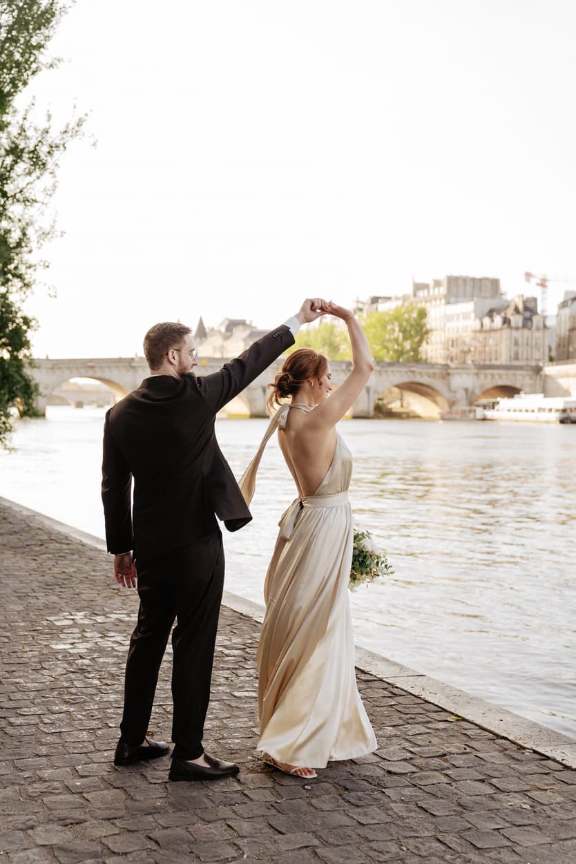 Bride and groom dance on a cobblestone Seine riverbank beside a bridge in warm evening light