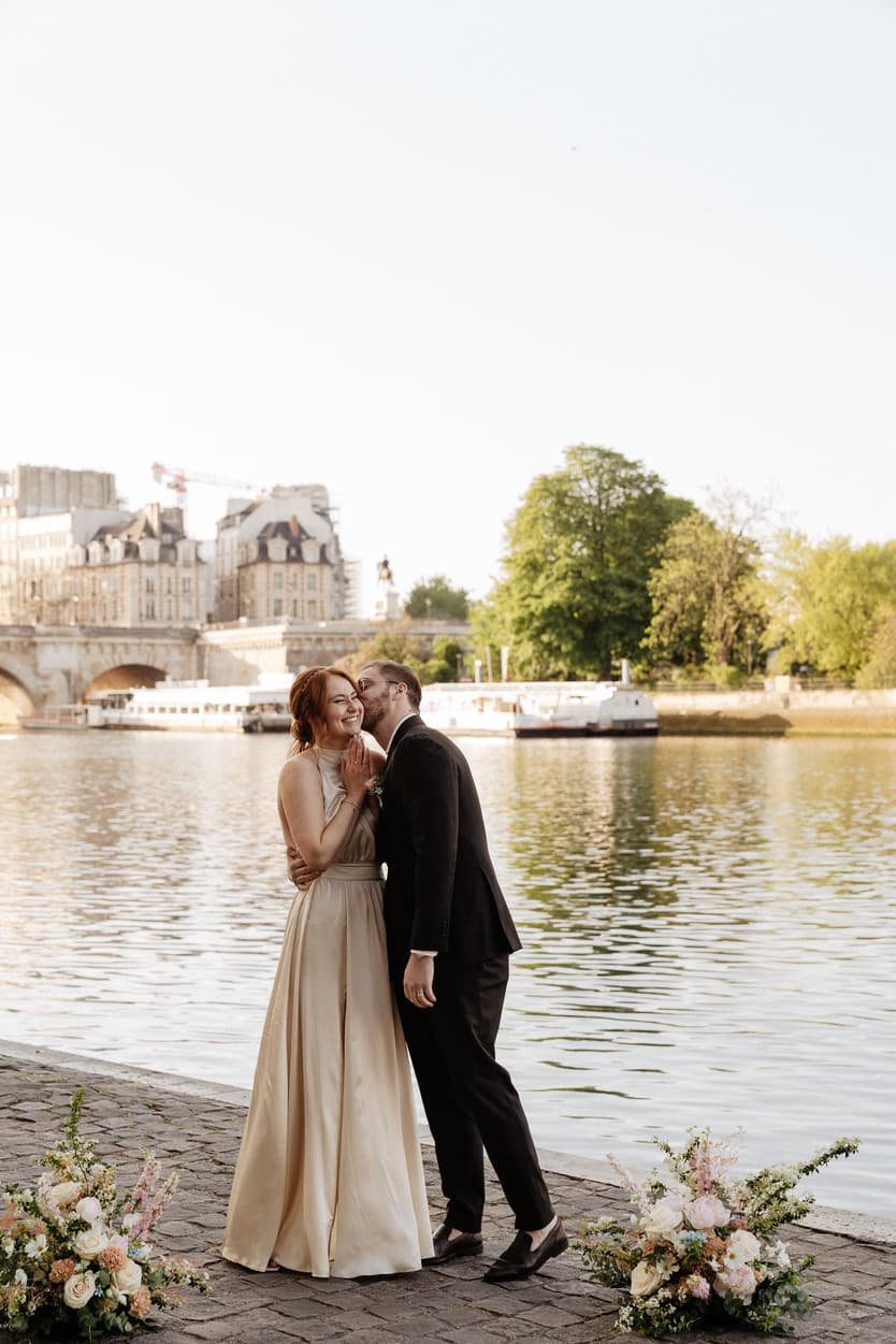 A bride and groom kiss on a Paris riverside at golden hour, framed by floral arrangements