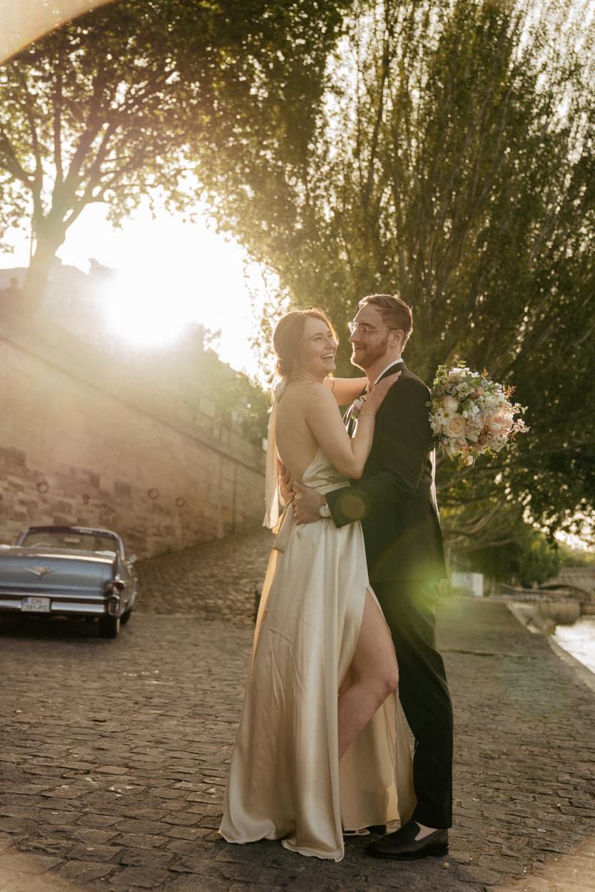 Bride and groom embrace on a Paris street at golden hour, bouquet in hand, beside a vintage car