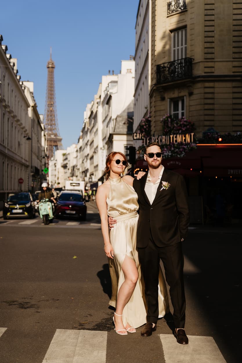Couple in formalwear pose on a Paris street with the Eiffel Tower behind them in warm afternoon light