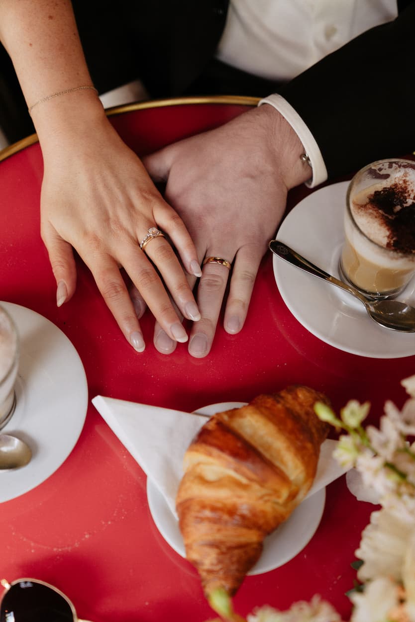 Bride and groom hold hands over a red café table with coffee, a croissant, and soft flowers