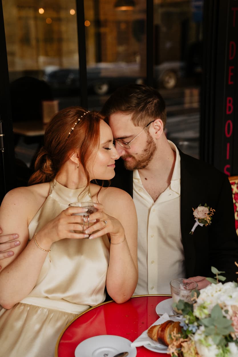 Bride and groom lean close at a Paris café, holding drinks by a red table with flowers