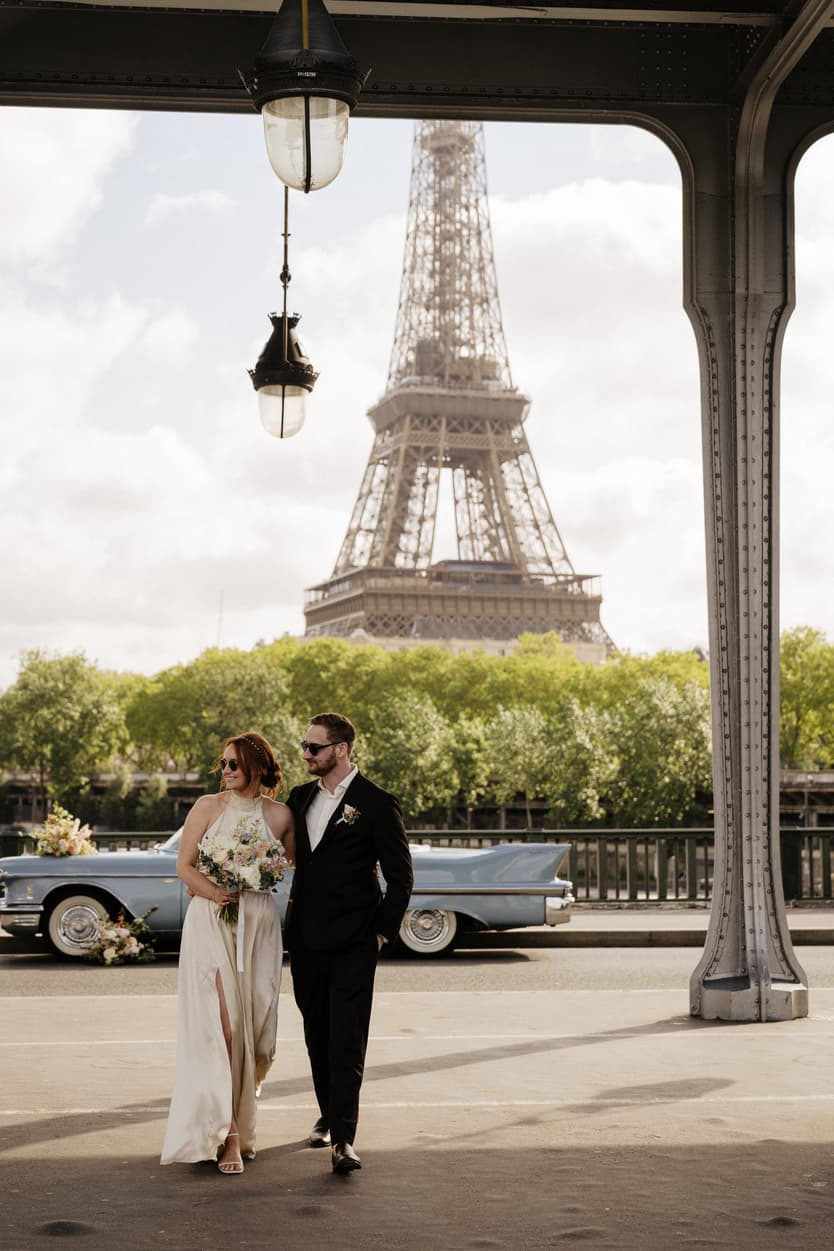 Bride and groom stand under a pavilion with the Eiffel Tower behind them and a vintage blue car nearby