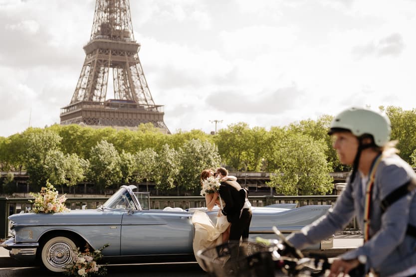 Bride and groom kiss beside a vintage car with the Eiffel Tower behind them as a cyclist passes by