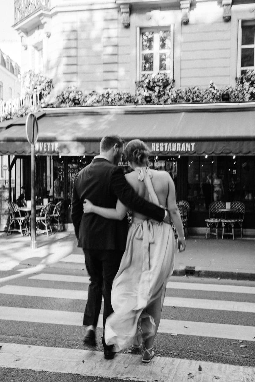Couple in wedding attire walks arm in arm across a Paris street in soft black-and-white light