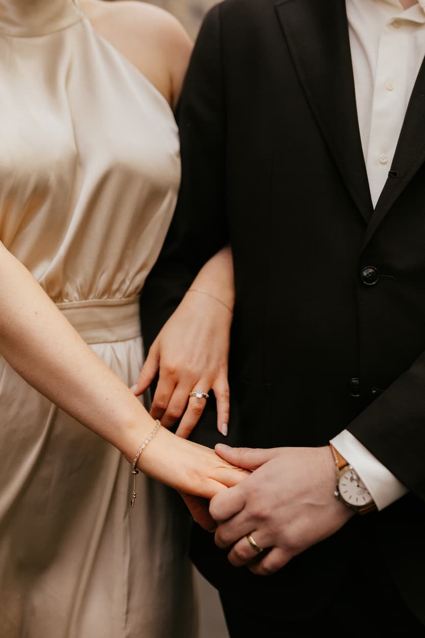 Bride and groom clasp hands in soft light, showing rings and a gold watch