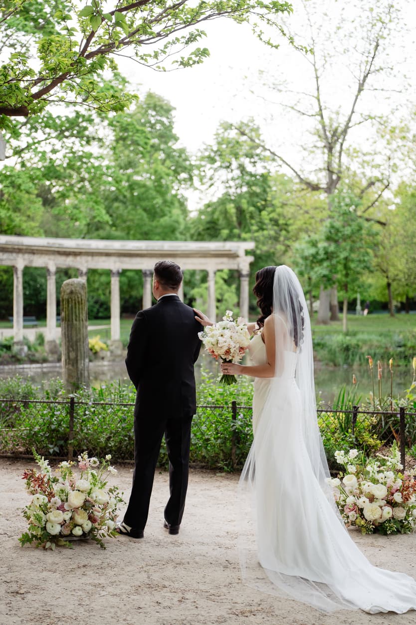 Bride in veil touches groom in black suit at a garden pond, framed by white floral arrangements and soft daylight