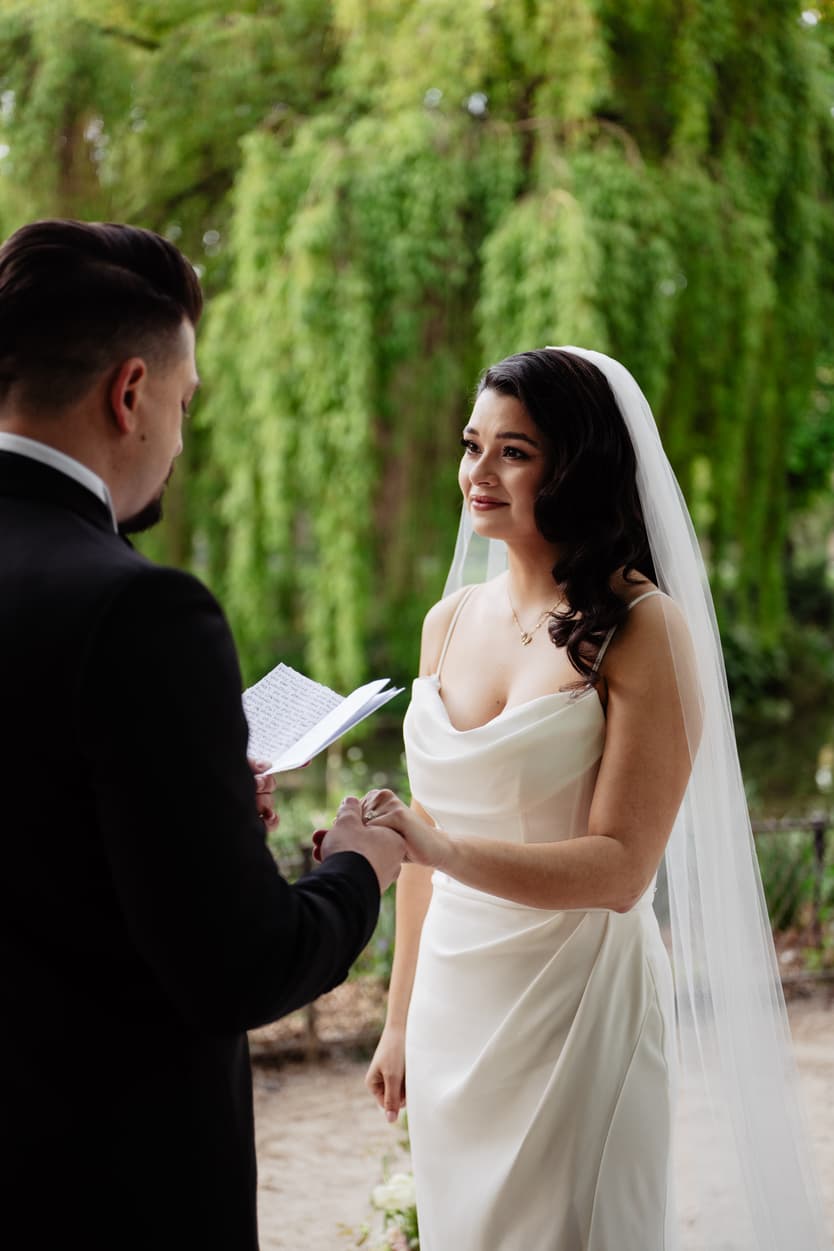 Bride and groom exchange rings in a lush garden, with a handwritten vow card and soft light