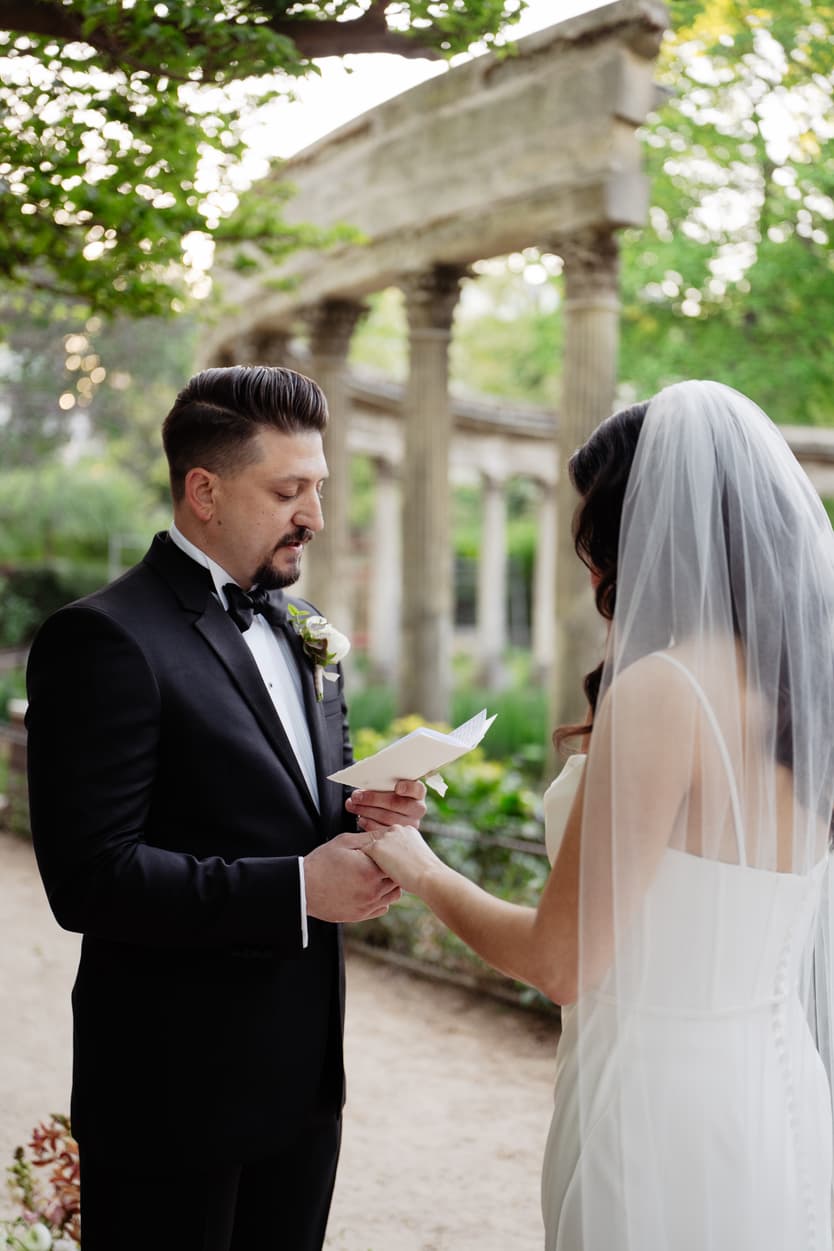 Bride and groom exchange vows in a sunlit garden, holding hands beneath stone columns and greenery