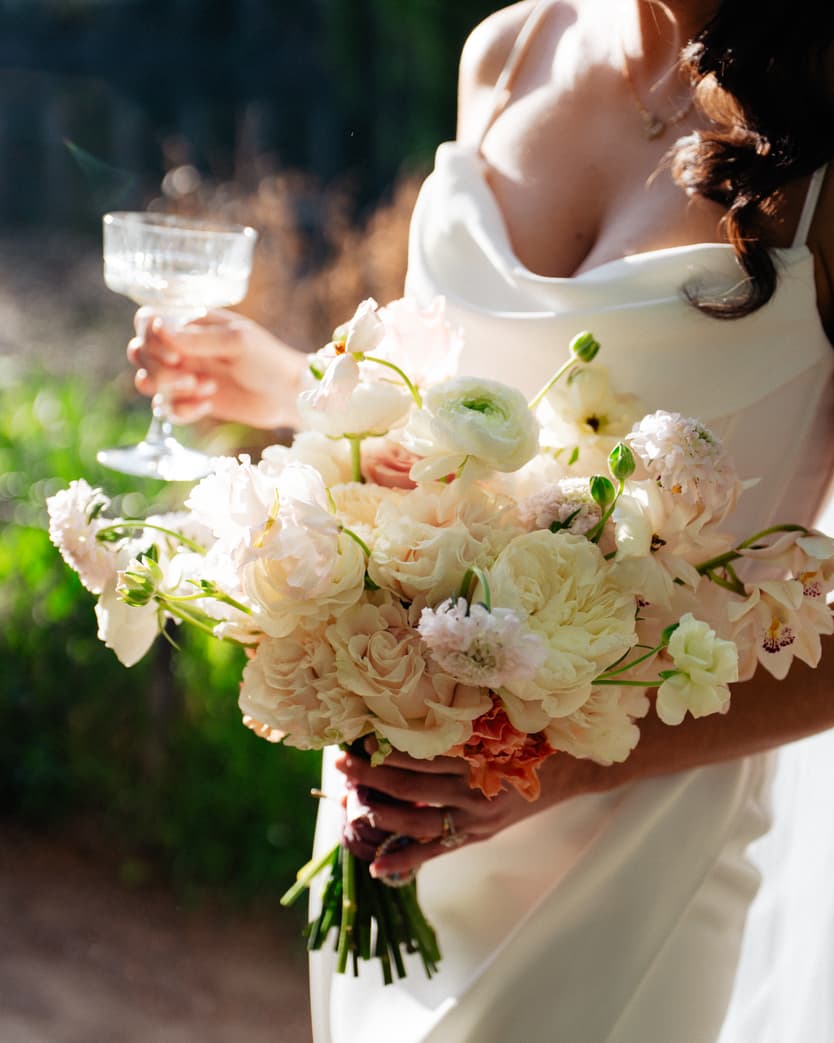 Bride in a white dress holds a pastel bouquet and champagne glass in a sunlit garden