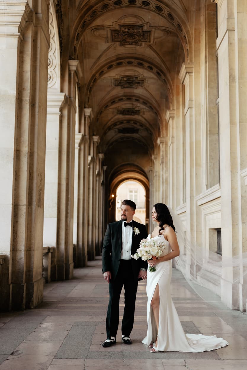 Bride and groom stand beneath a grand Paris arcade, holding a bouquet in warm light