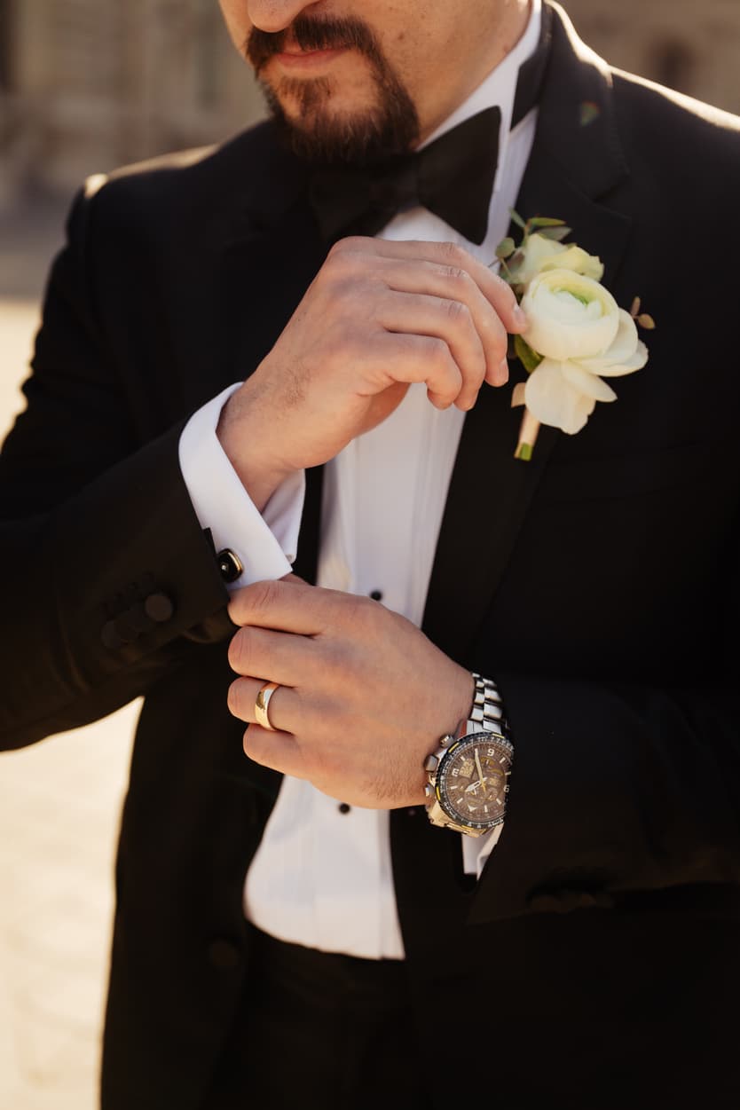 Groom in a black tuxedo adjusts a white boutonniere in warm light on a Paris street