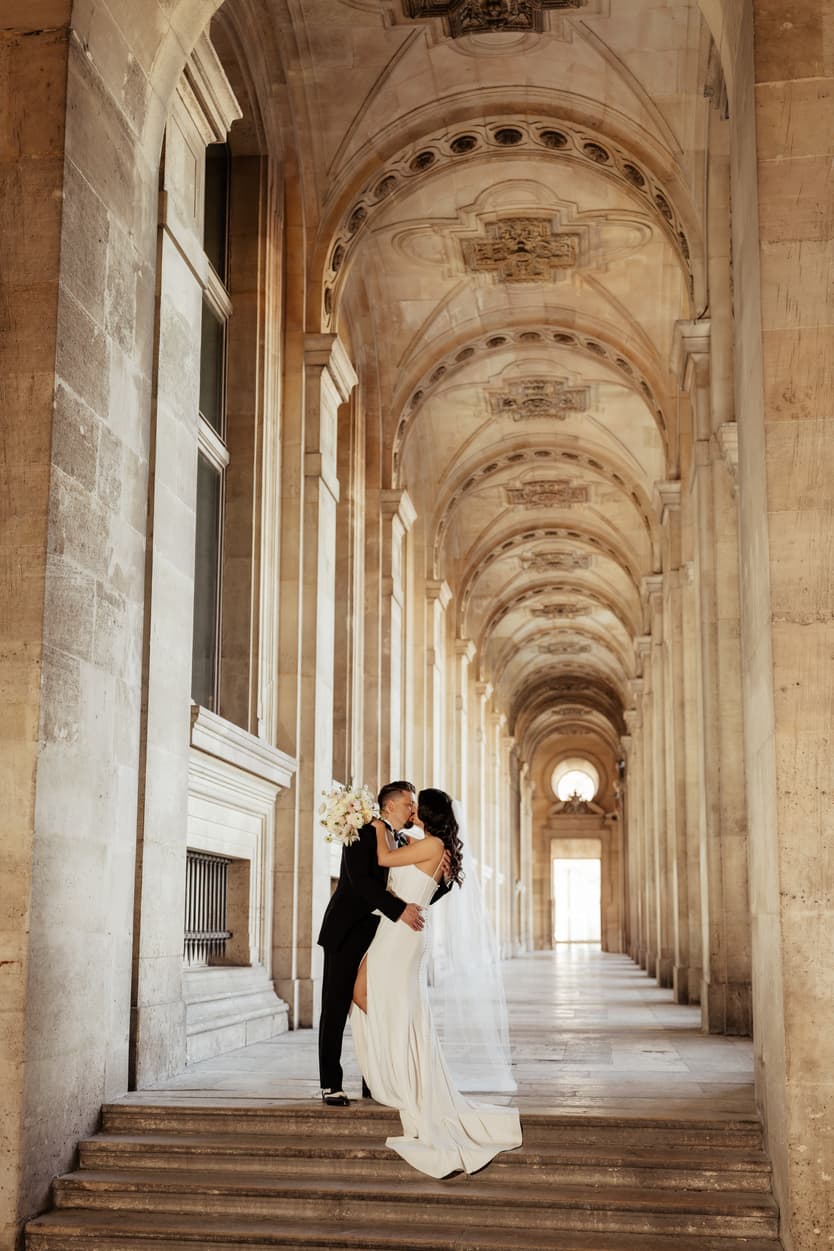 Bride and groom kiss on palace steps in warm light beneath a vaulted Paris arcade