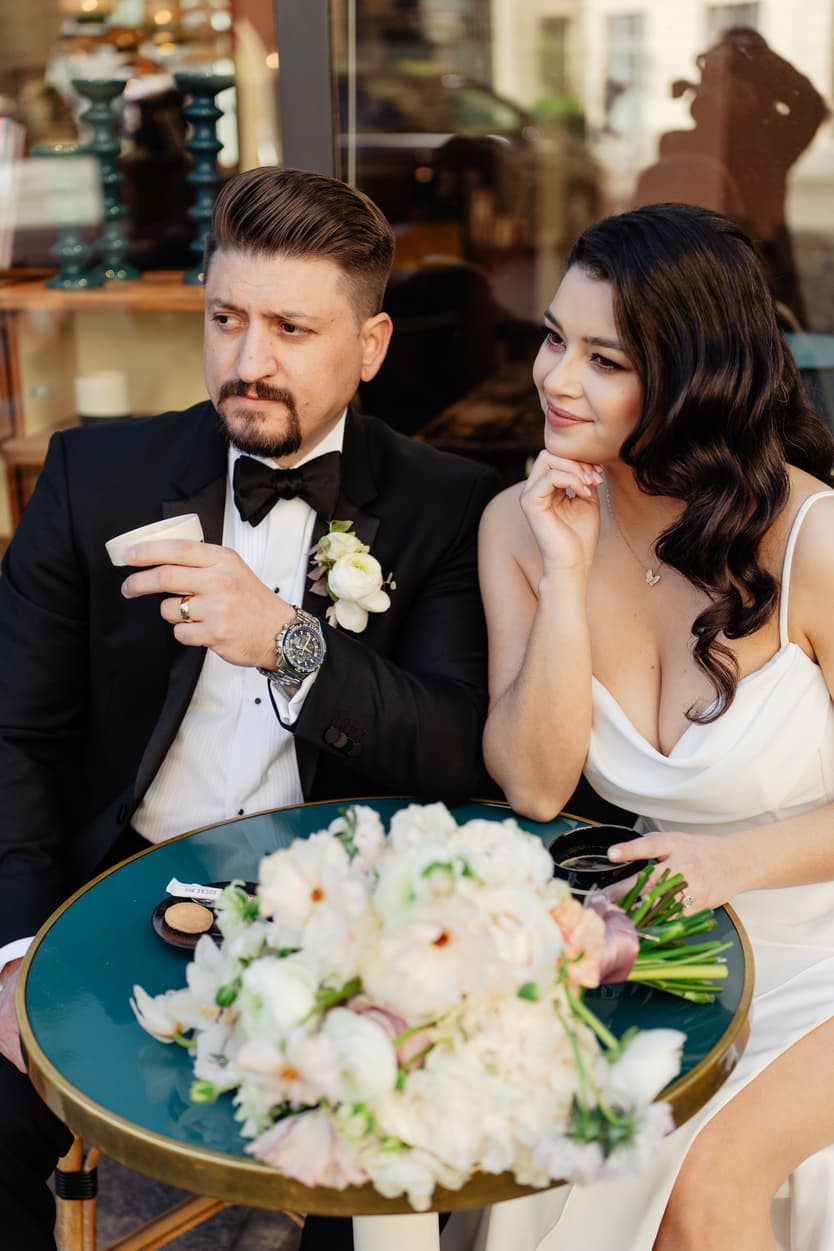 A bride and groom at a green café table in Paris, her bouquet of white and peach roses beside them