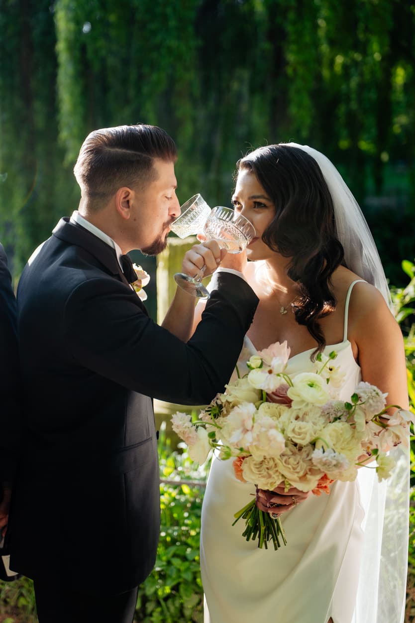 Bride and groom sip from crystal goblets in a sunlit garden, bouquet in hand and veil trailing behind
