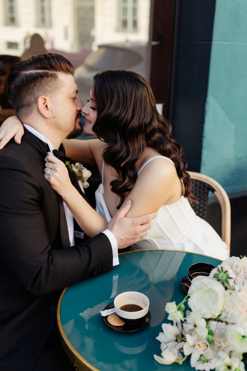 Bride and groom kiss at a Paris café table, sunlight on their faces and a bouquet beside coffee cups