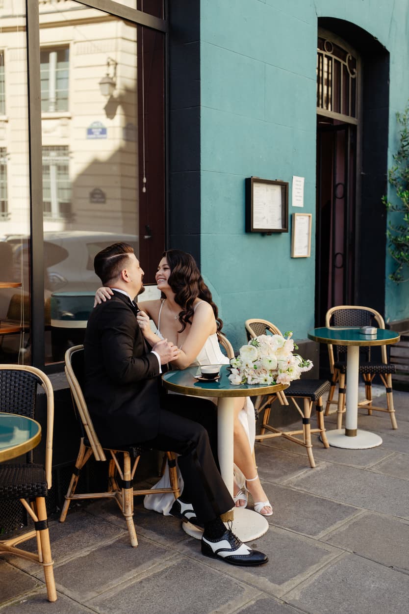 Bride and groom sit at a café table on a Paris street, smiling in soft daylight beside a bouquet