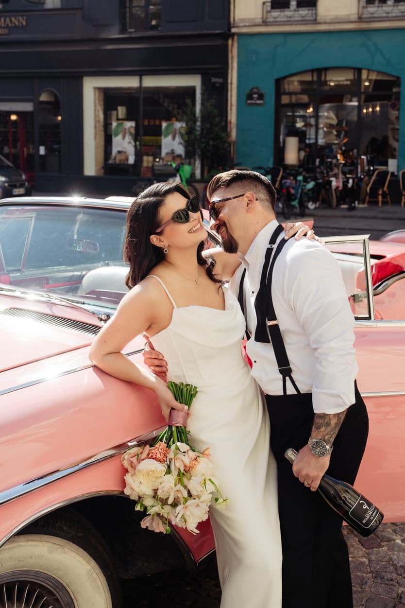Bride and groom pose beside a pink vintage car on a Paris street in warm sunlight, bouquet and champagne in hand