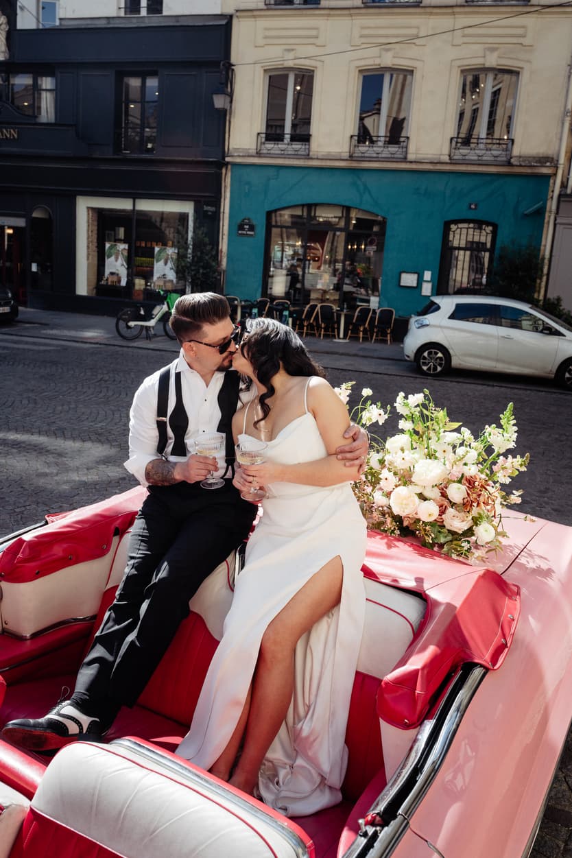 A bride and groom kiss in a pink convertible on a Paris street, with white flowers and bright sun