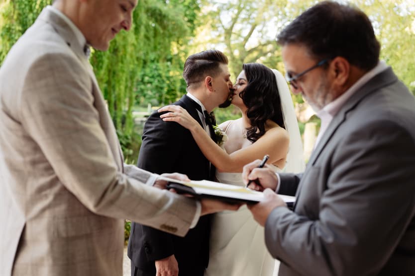 Bride and groom kiss as an officiant signs papers, with a guest nearby in a green garden