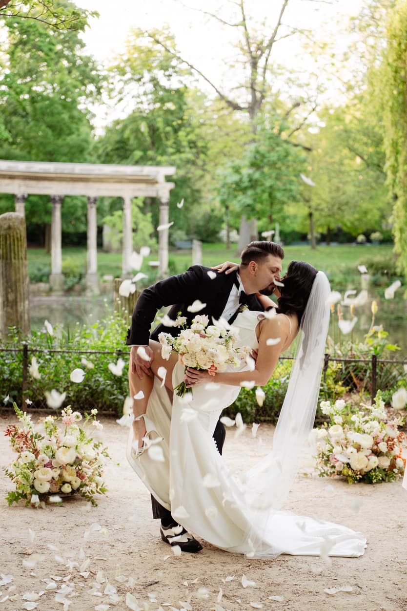 Bride and groom kiss in a garden dip beside a pond, with petals falling and floral arrangements