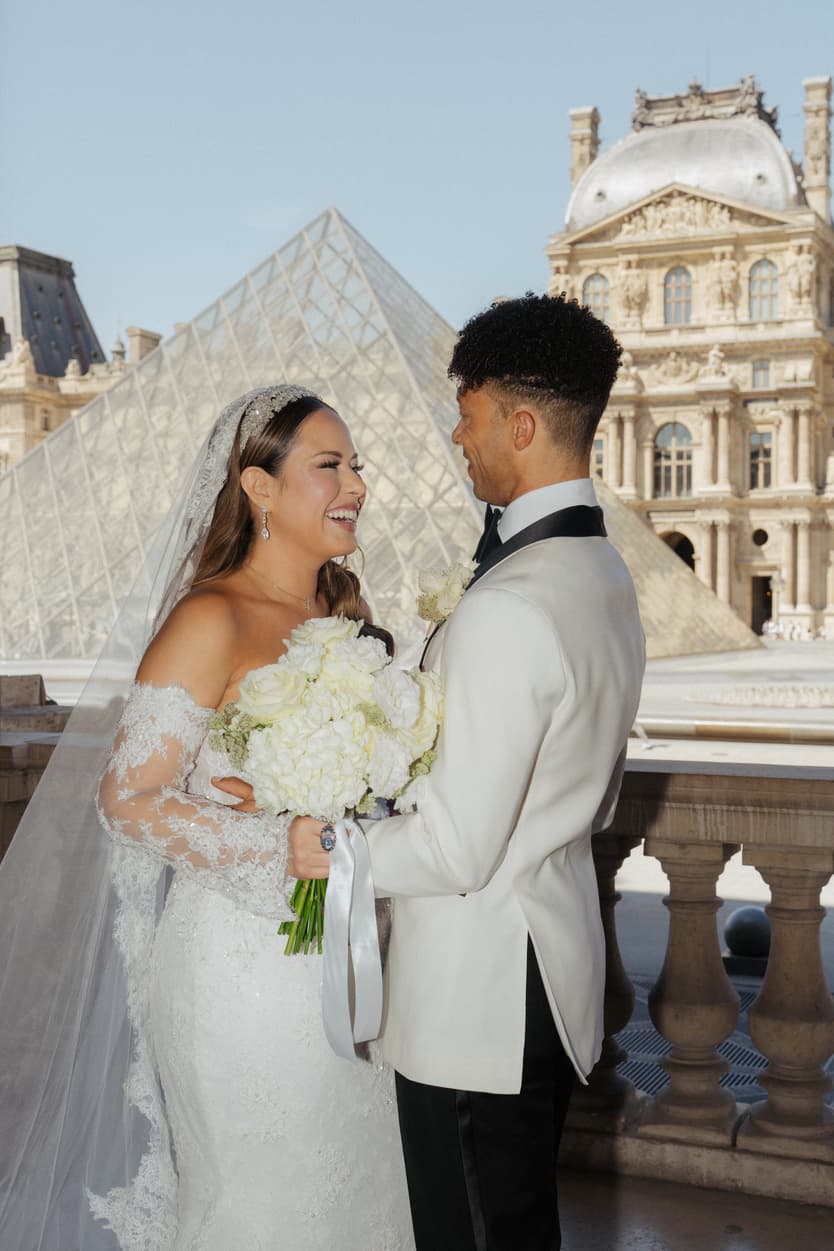 Bride and groom smile by the Louvre pyramid, holding white flowers on a sunlit terrace