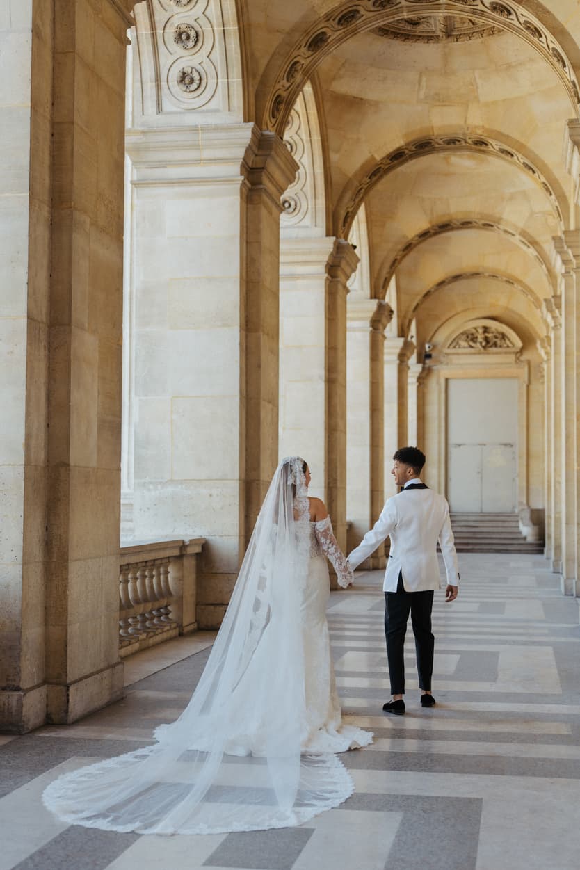 Bride and groom walk hand in hand through a sunlit arched colonnade, her veil trailing behind