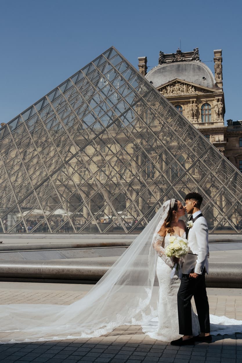 Bride and groom kiss before the Louvre pyramid, her veil trailing across the sunlit plaza