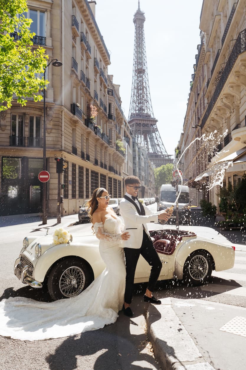 Bride and groom pop champagne beside a vintage car on a Paris street with the Eiffel Tower behind
