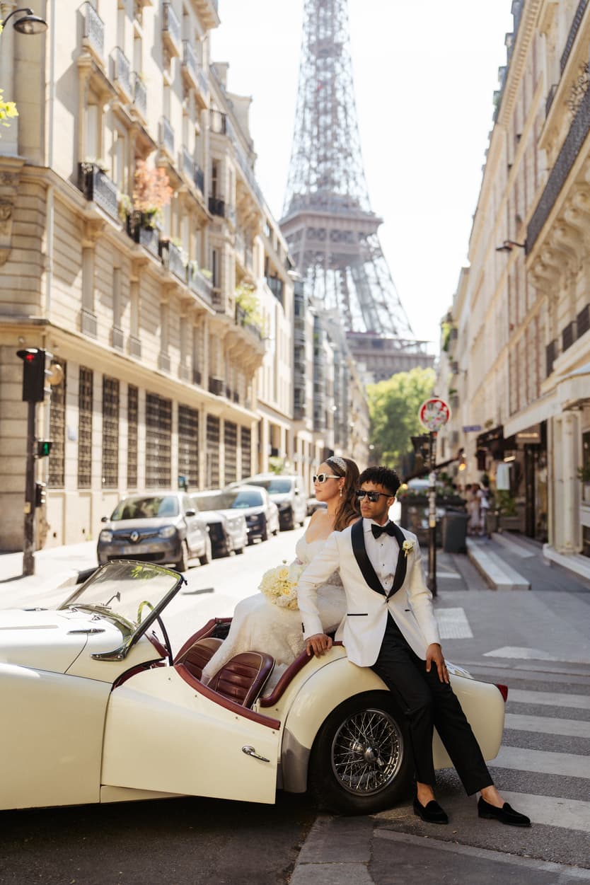 Bride and groom pose on a Paris street in bright light, with the Eiffel Tower rising behind them