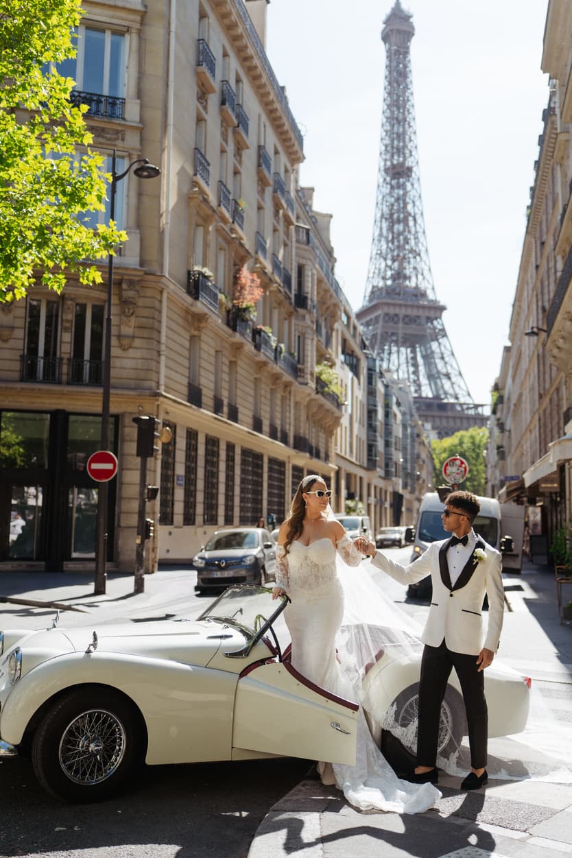 Bride and groom pose by a vintage car on a Paris street with the Eiffel Tower behind them
