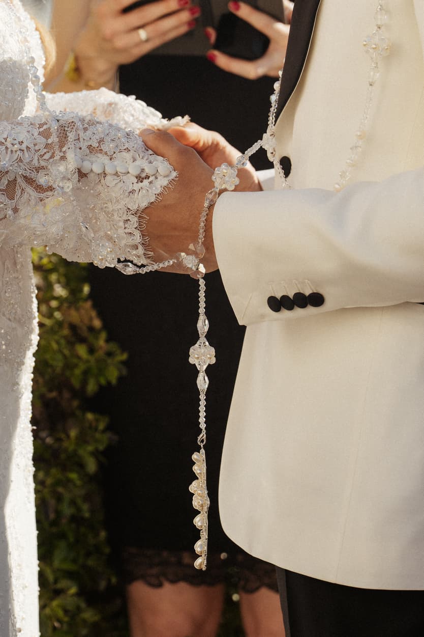 Bride and groom clasp hands in warm light, her lace sleeve and crystal tassel against his white tuxedo