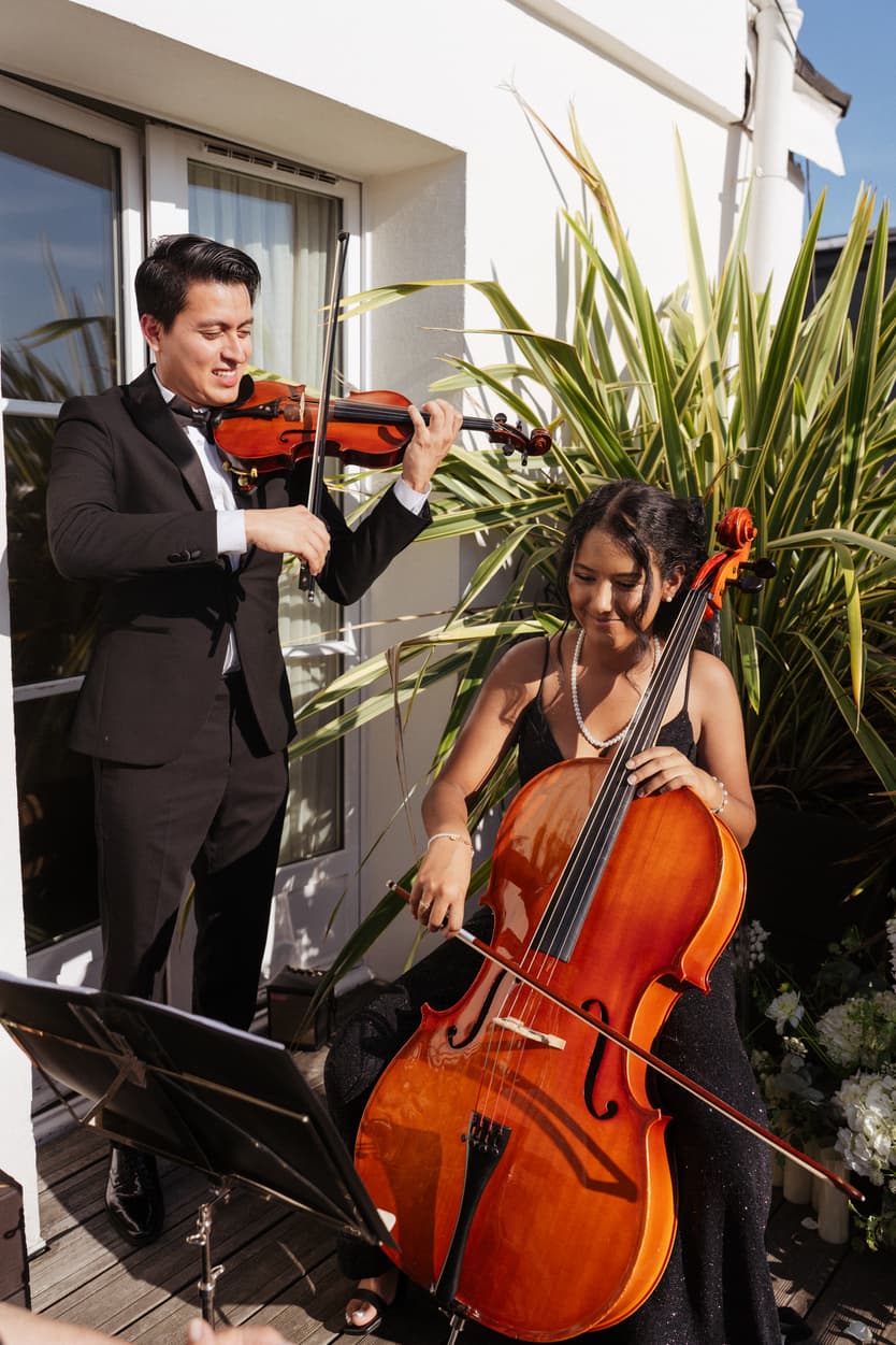 A violinist and cellist play on a sunlit terrace beside a white facade and potted greenery