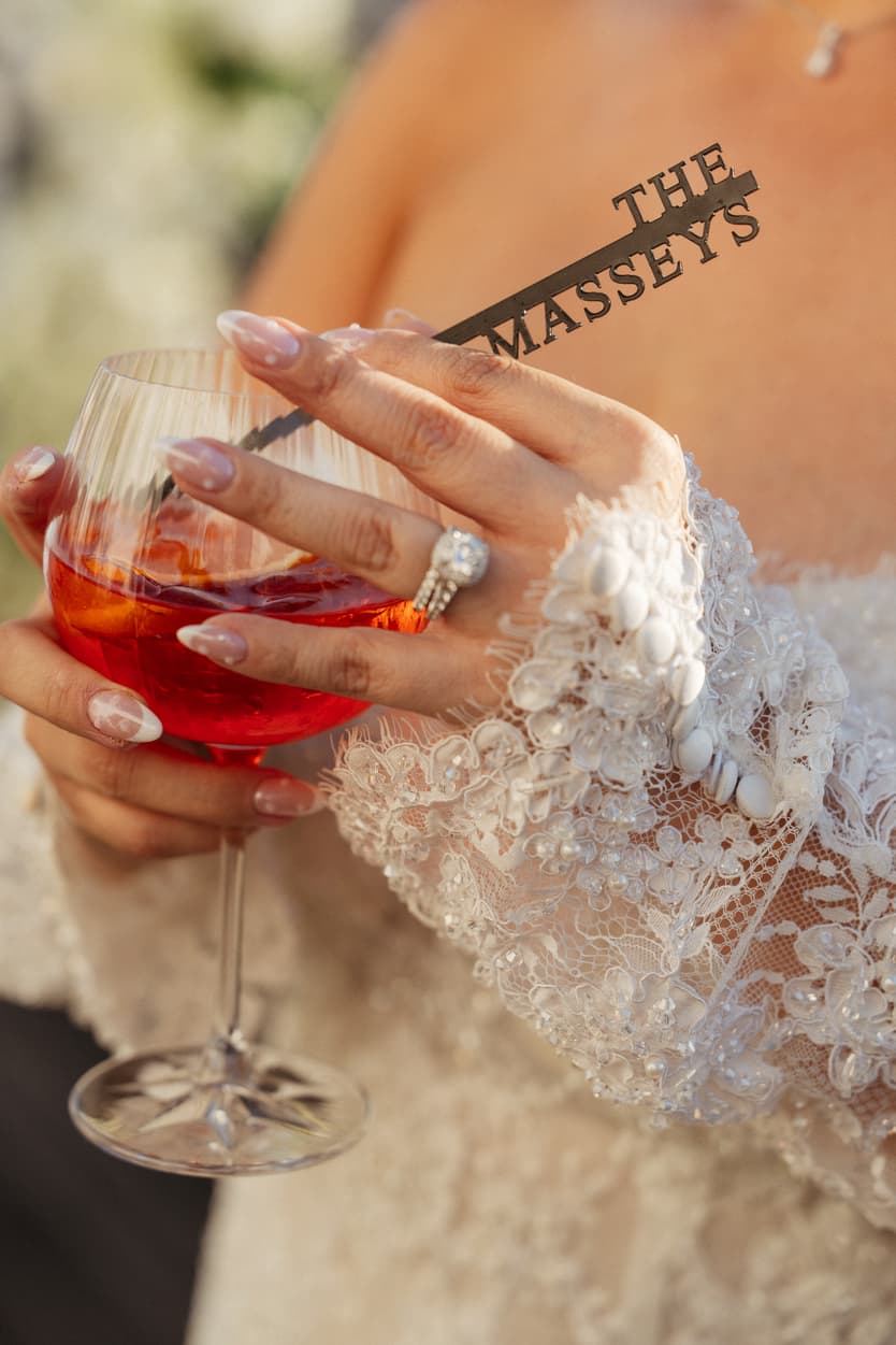 Bride in a lace sleeve holds a red cocktail with a THE MASSEYS stirrer in warm light