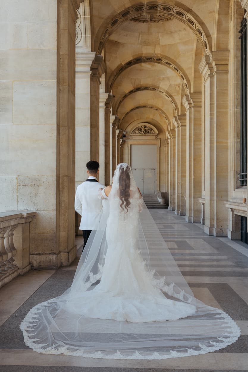 Bride and groom walk through a sunlit stone arcade, her long veil trailing behind