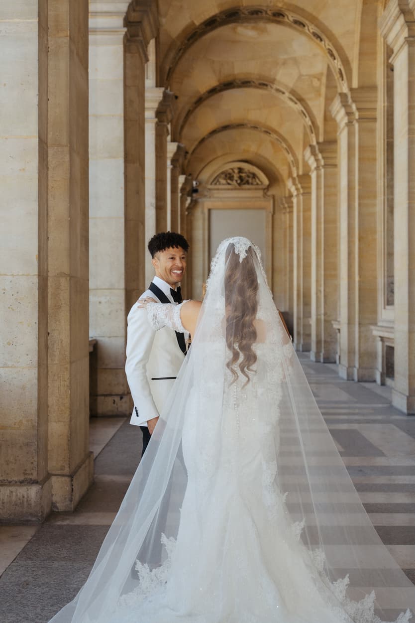 Bride and groom share a smile in a sunlit Paris arcade with stone arches and her long veil