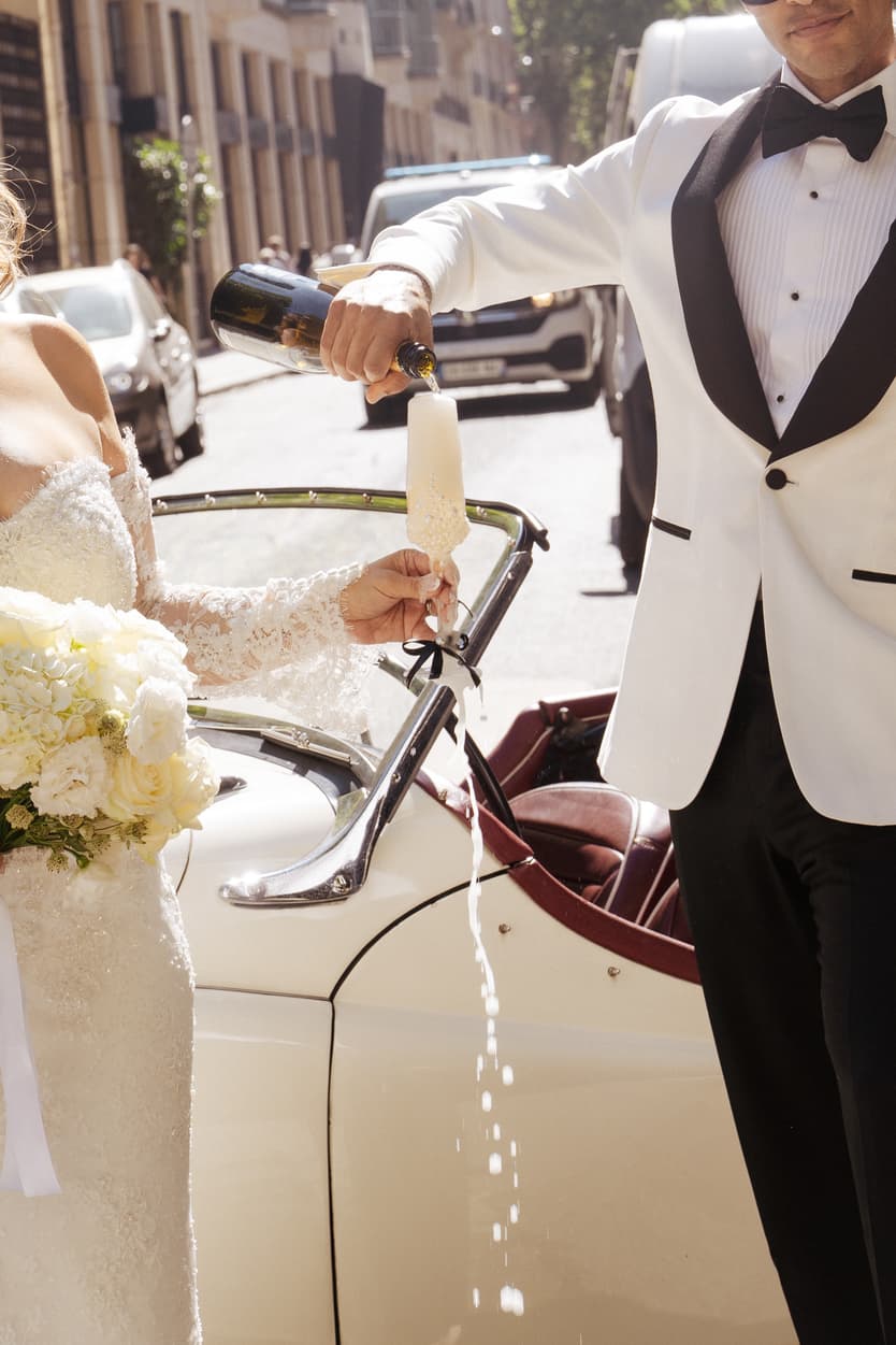 Bride and groom toast champagne beside a vintage car on a sunny Paris street